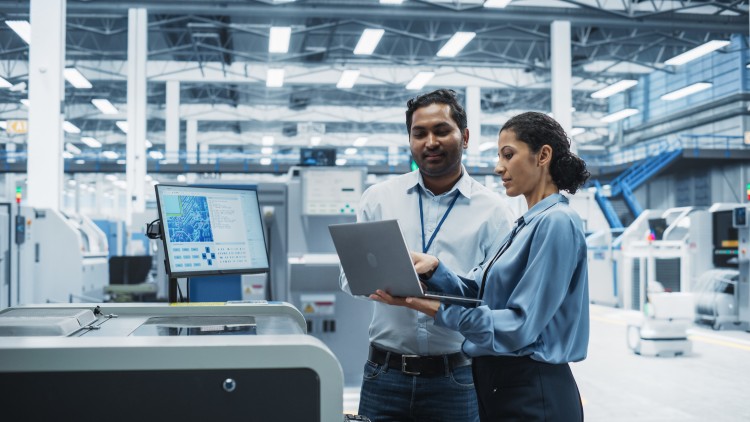 Two industrial engineering technicians look at a laptop on a factory floor.