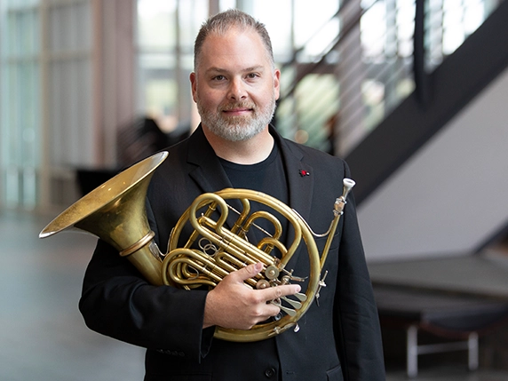 A man in a suit holds a french horn.